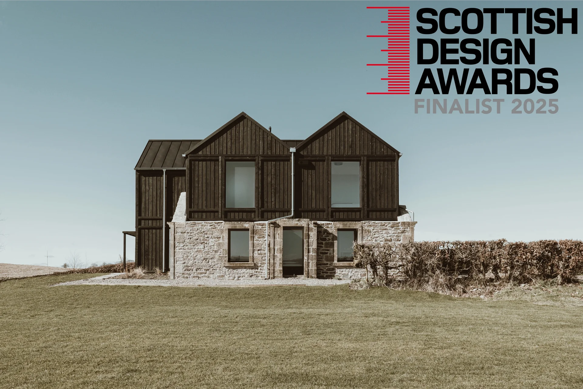 Modern rural home with dark timber cladding and stone ground floor, set in open countryside under a clear sky, with "Scottish Design Awards Finalist 2025" logo in the top-right corner
