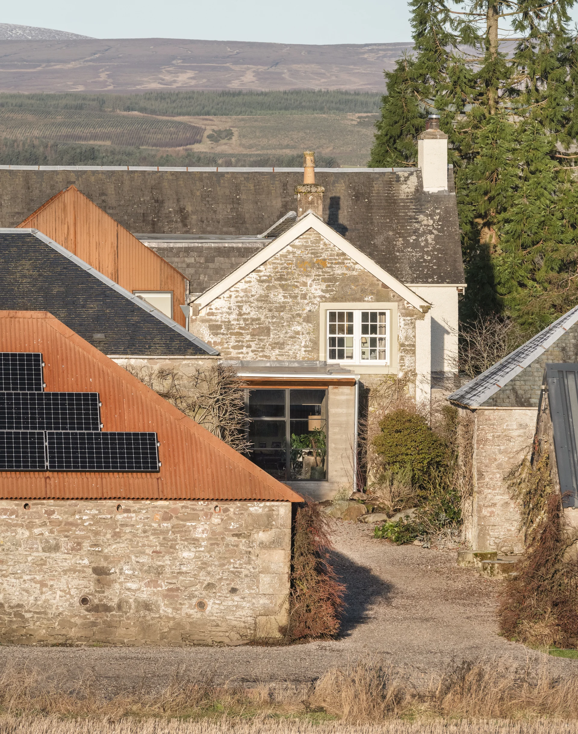 Stone farmhouse with a modern extension and solar panels on an adjacent outbuilding, blending traditional architecture with renewable energy.