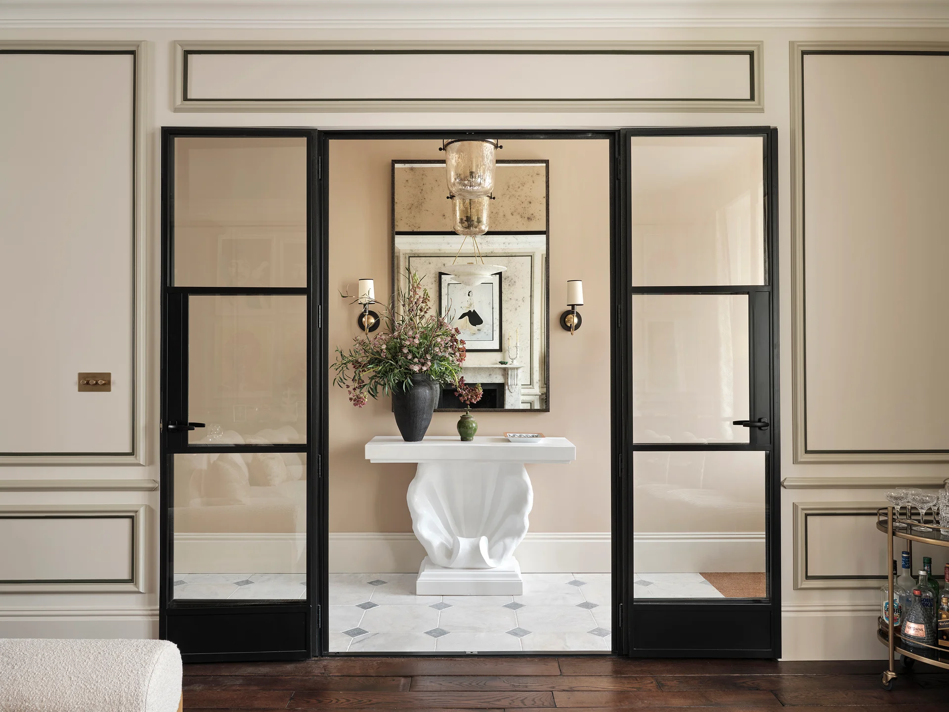 Elegant interior hallway with black-framed glass doors, a sculptural white console table, large mirror, and decorative wall sconces.