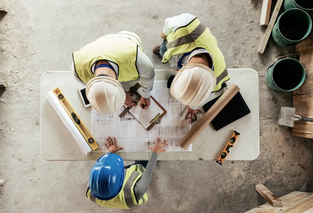Overhead view of three construction workers in safety vests and helmets reviewing architectural plans on a site table.
