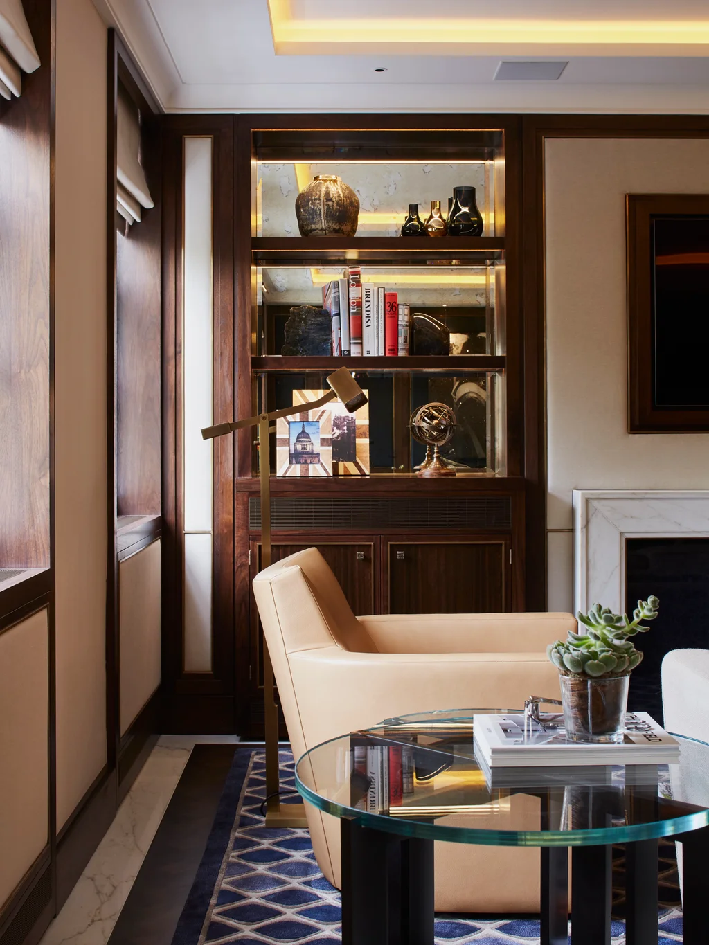 Stylish living room corner with a leather armchair, glass coffee table, and illuminated shelving displaying books and decorative objects.