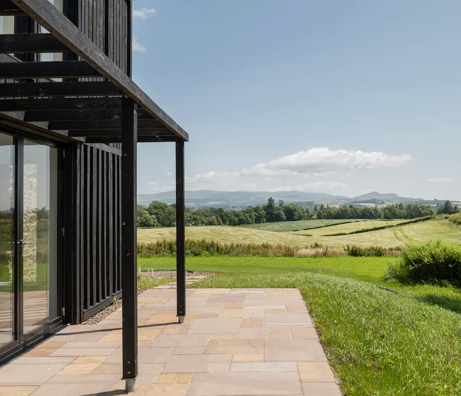 View from a modern black timber and stone house onto a stone patio overlooking rolling green fields and distant hills.