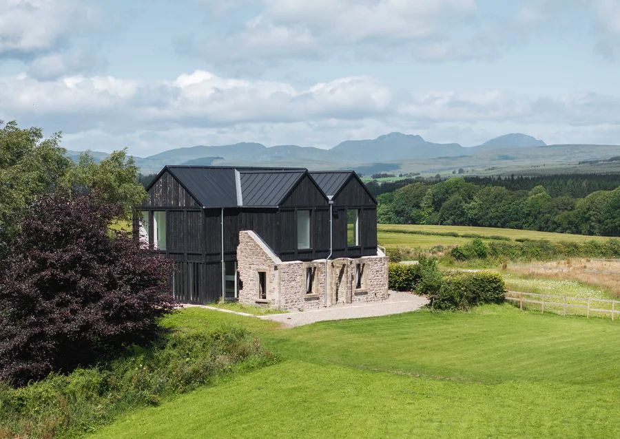 Contemporary black timber and stone-clad house set in the countryside with mountains and farmland in the background.