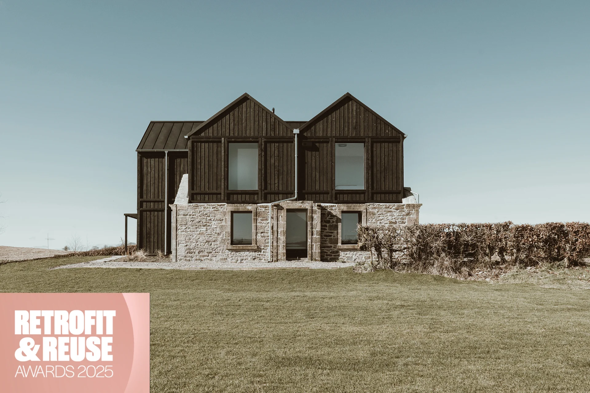 Front view of a black timber and stone house with large windows, shown with a Retrofit & Reuse Awards 2025 badge.