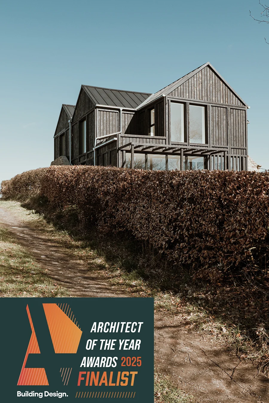 Angled view of a dark timber-clad house with expansive glazing, seen behind a hedge with an Architect of the Year Awards 2025 Finalist graphic.