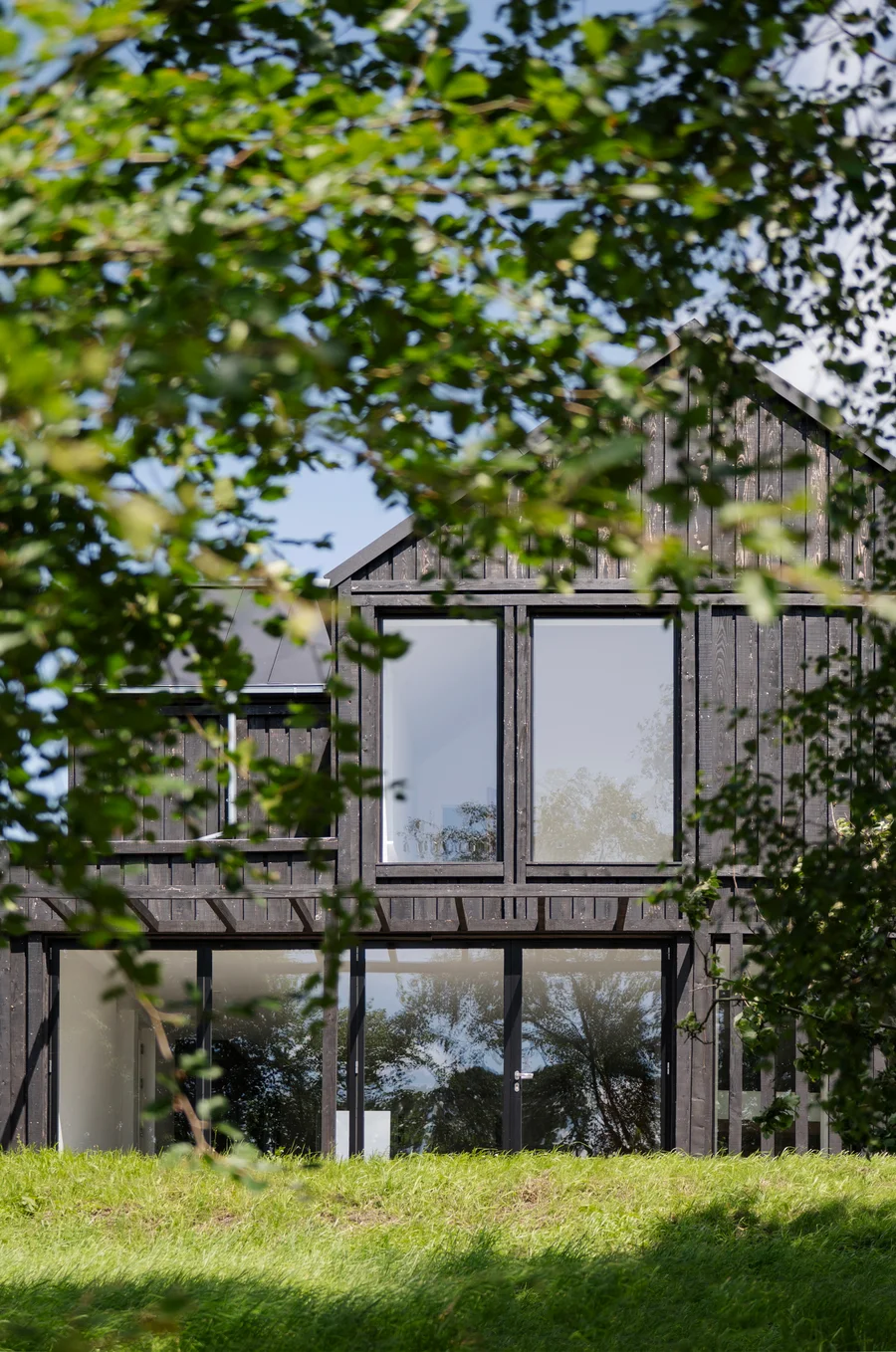 Black timber house partially hidden behind leafy green trees, showcasing its tall windows and modern barn-style design.