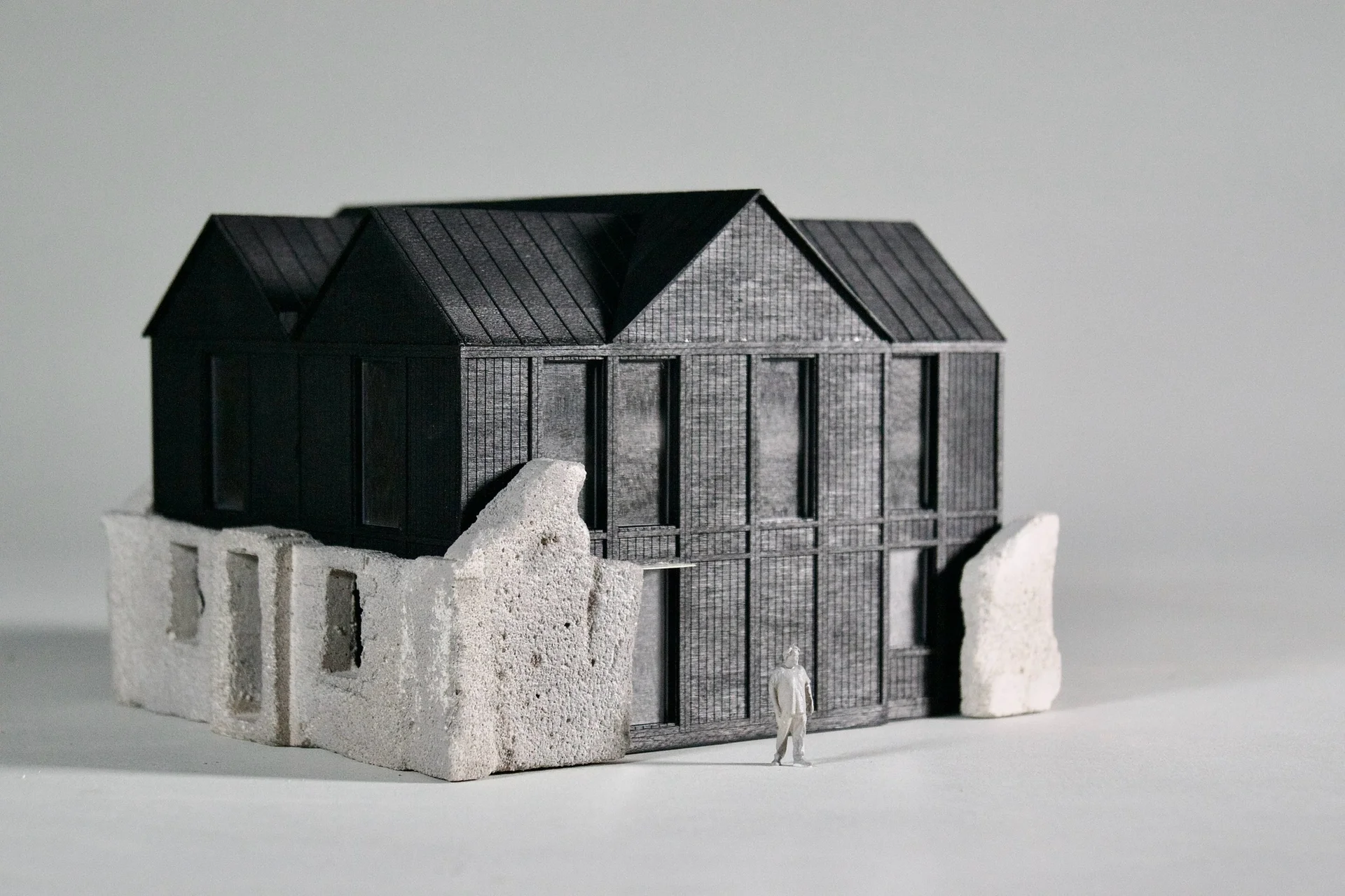 Architectural model of a house with dark timber cladding built onto the ruins of a stone structure, with a small human figure for scale.