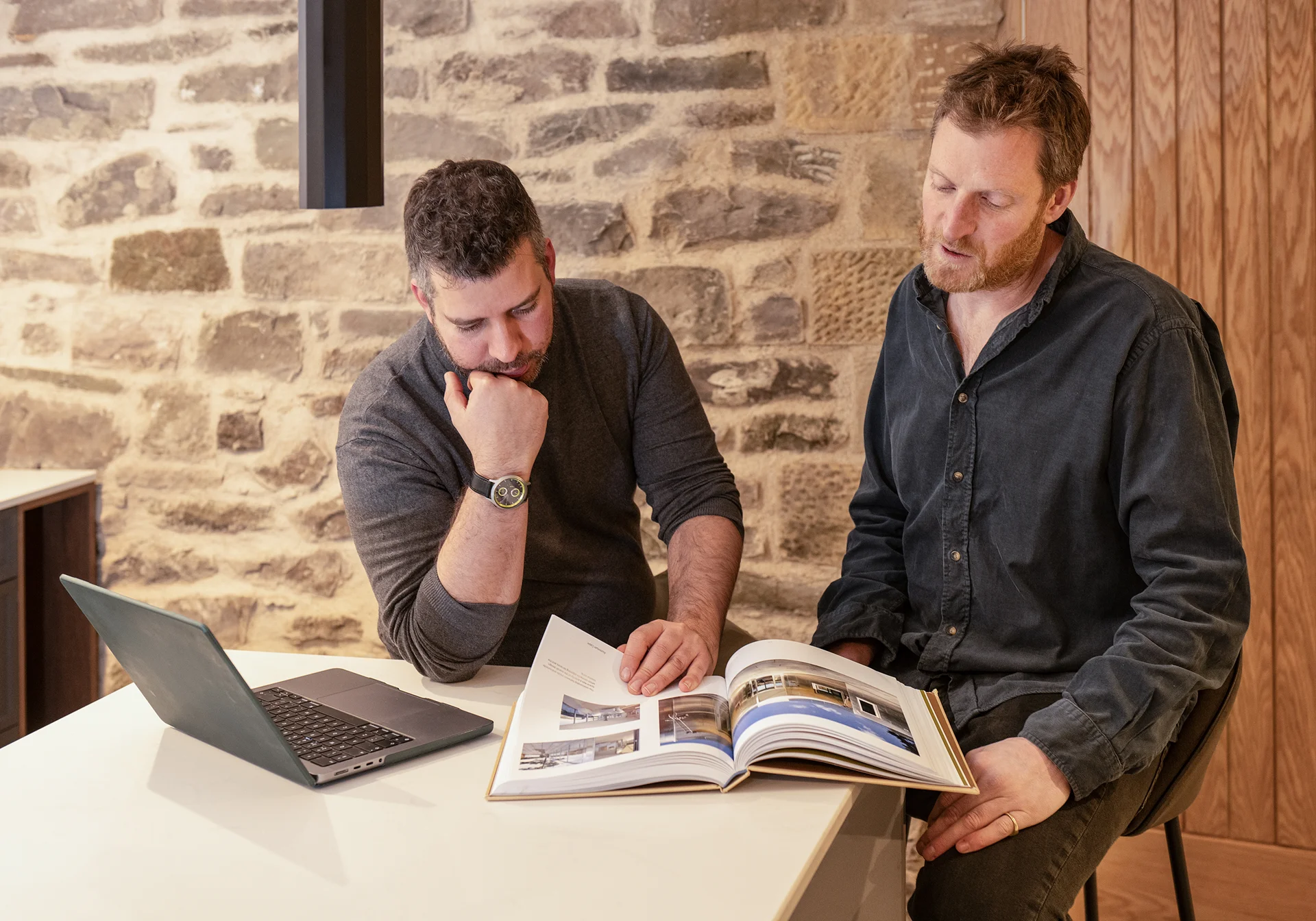 Two men sitting at a kitchen table discussing a project while looking at an open architecture book beside a laptop.