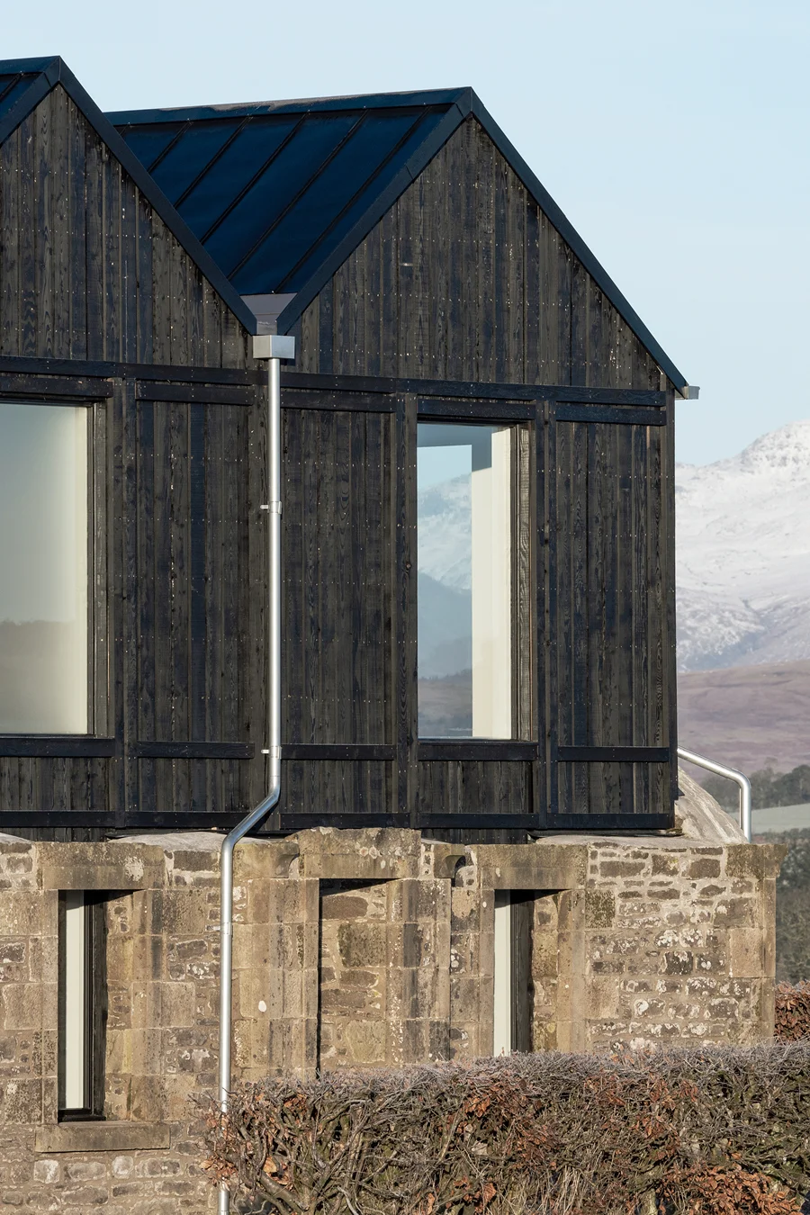 Modern black timber-clad extension built on top of an old stone building with mountains in the background.