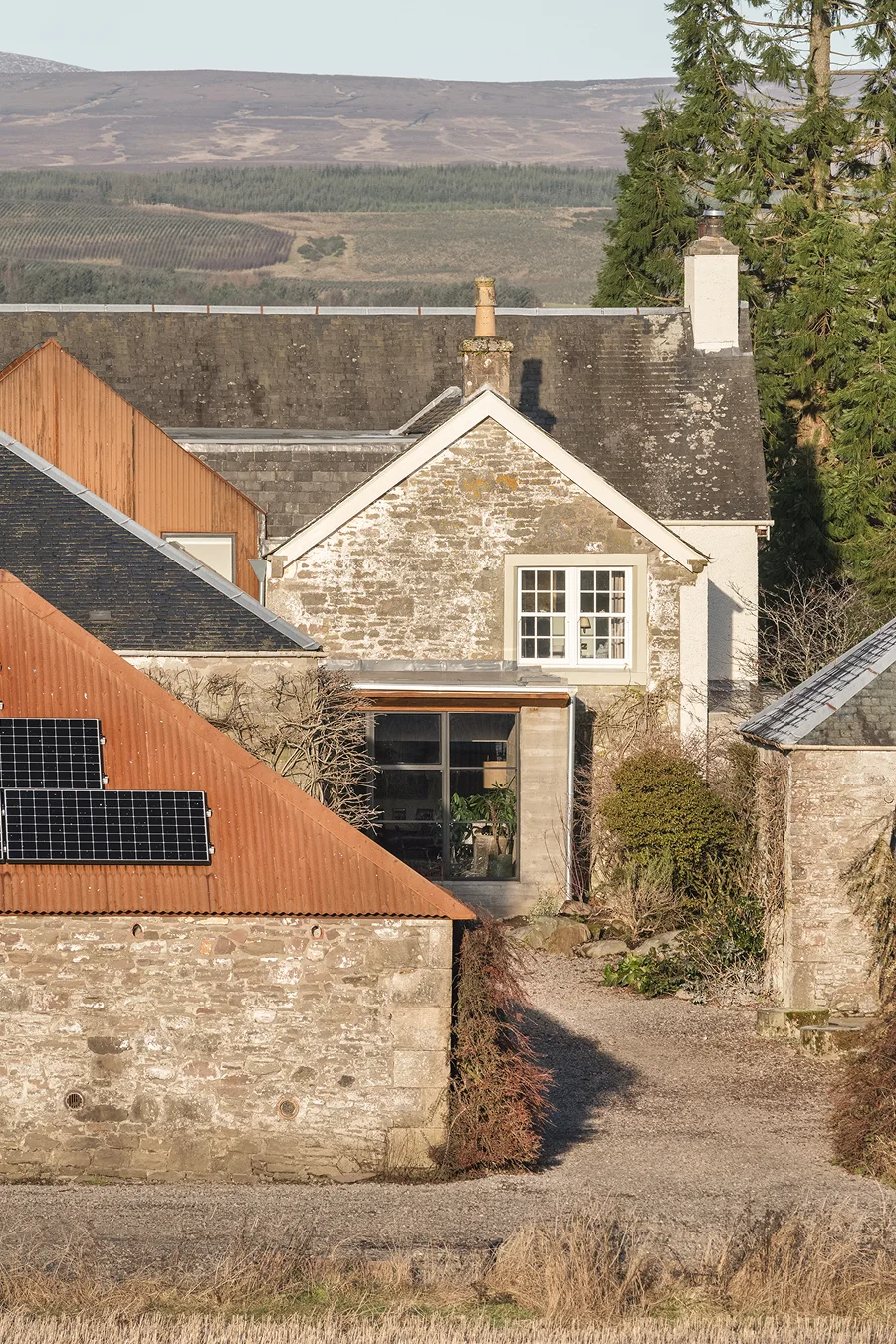 Restored countryside home with solar panels on an outbuilding and stone walls surrounded by trees and fields.