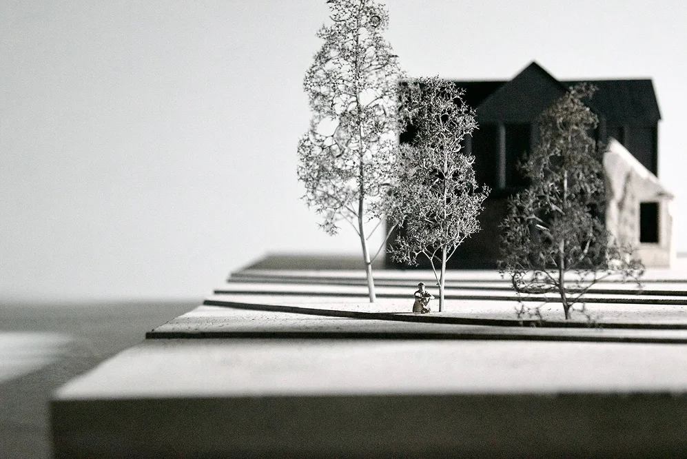Architectural model showing a dark house with trees and a small human figure in the foreground, designed on layered platforms.