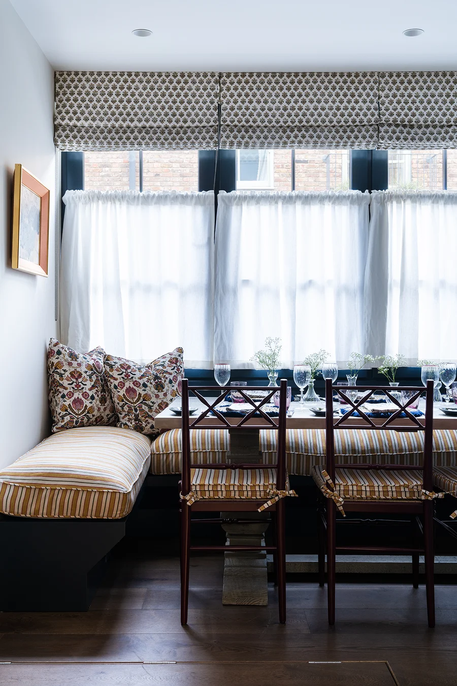 Charming dining nook with striped bench seating, patterned cushions, and a light-filled window dressed with roman blinds and café curtains.