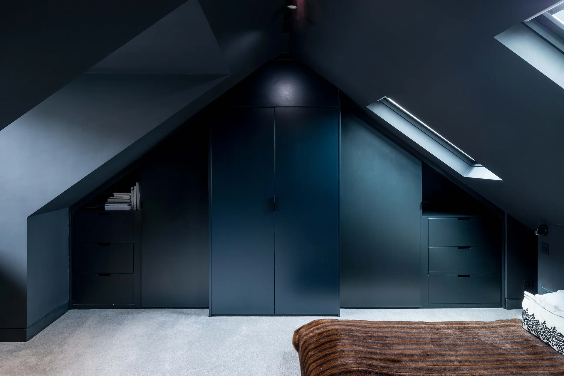 Moody attic bedroom with dark blue built-in wardrobes and skylights allowing natural light into the space.