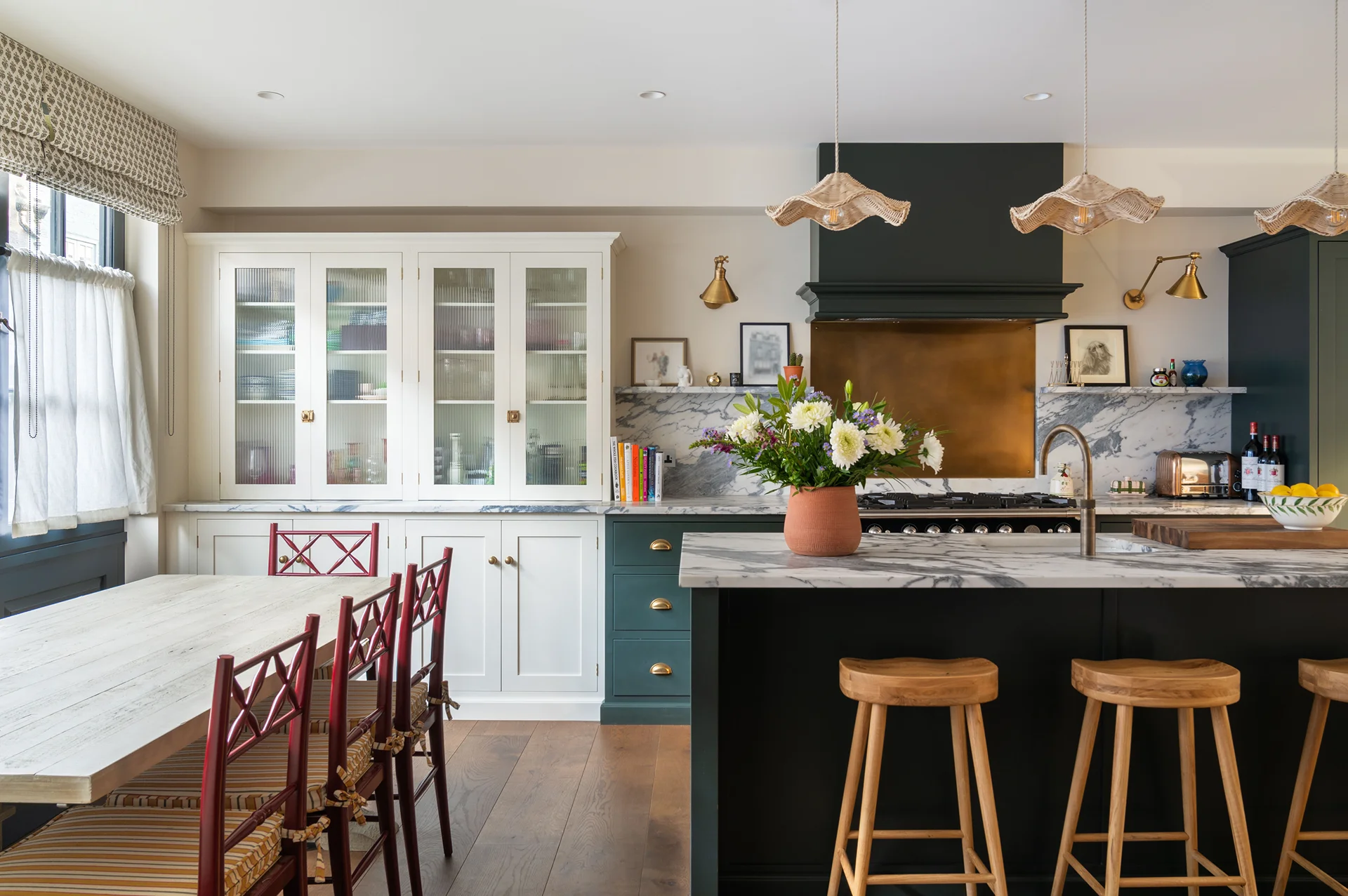 Stylish open-plan kitchen with marble worktops, brass accents, green cabinetry, and wooden stools beside a dining area with red chairs.