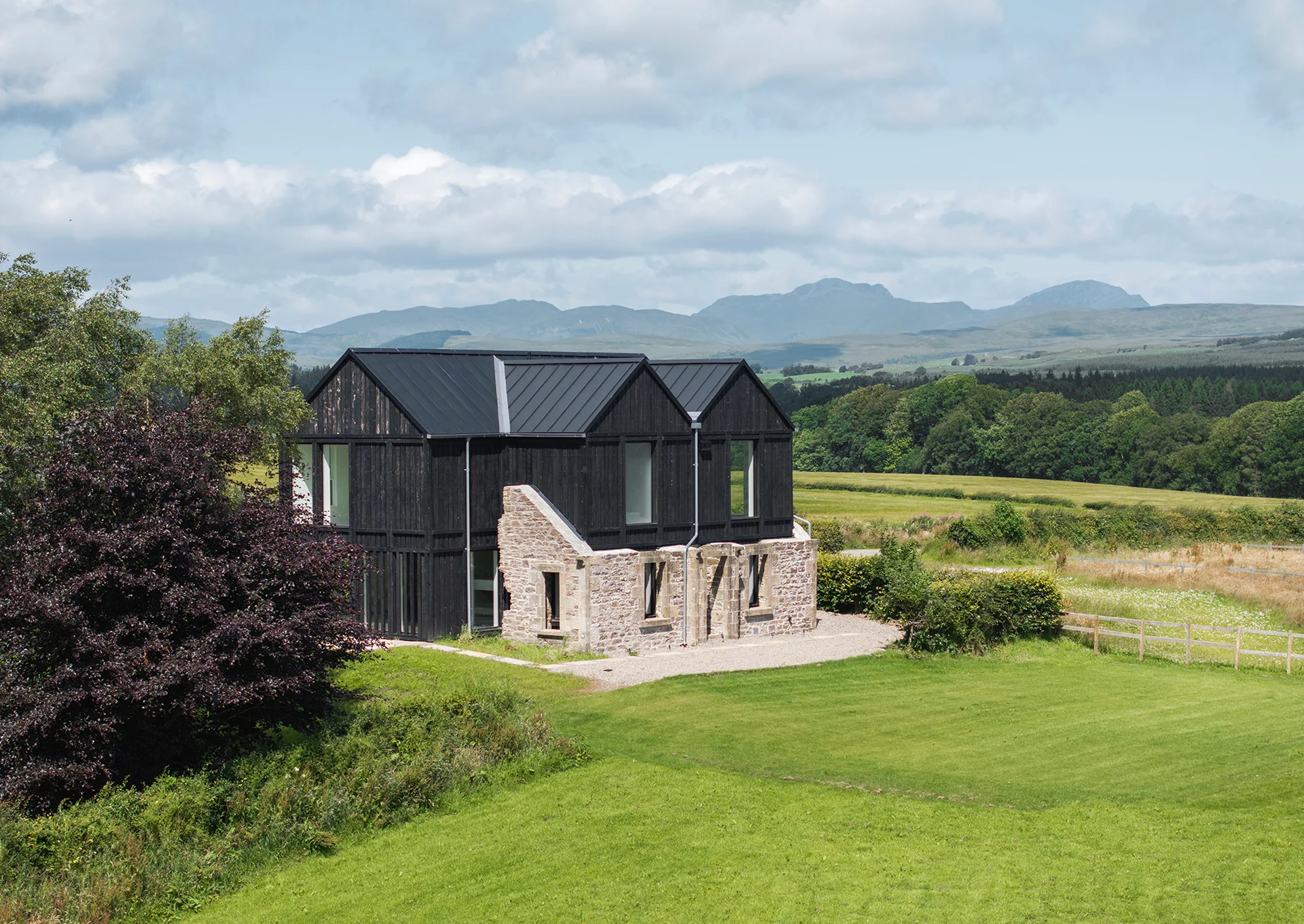 Modern black and stone house set in lush green landscape with mountains in the background under a partly cloudy sky.