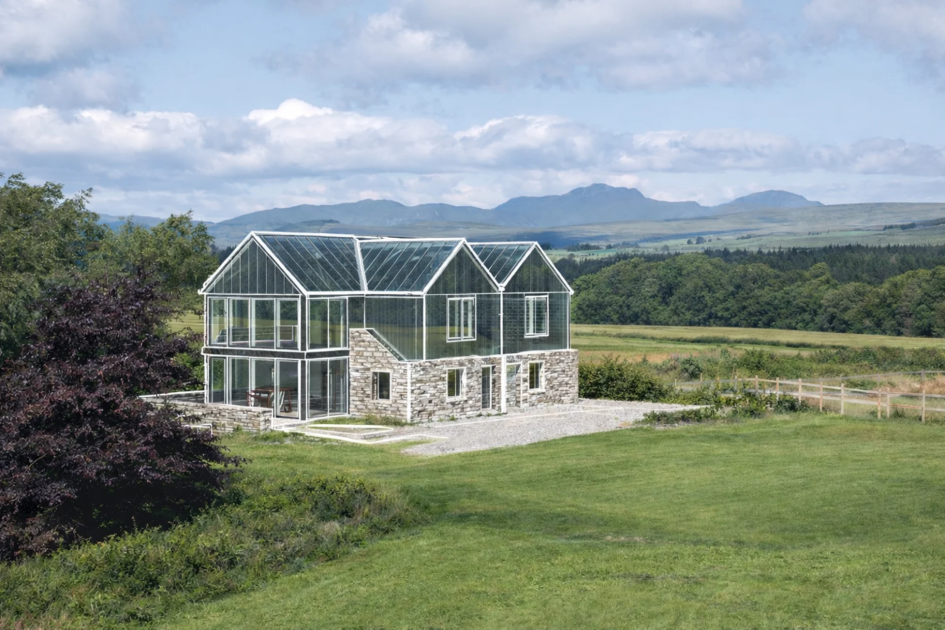 Modern glass house surrounded by greenery and mountains under a blue sky.