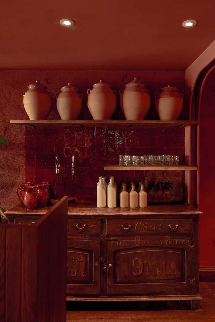 Wooden counter with stacked clay pots and bottles, set against a warm, red backdrop. Soft lighting highlights the scene.