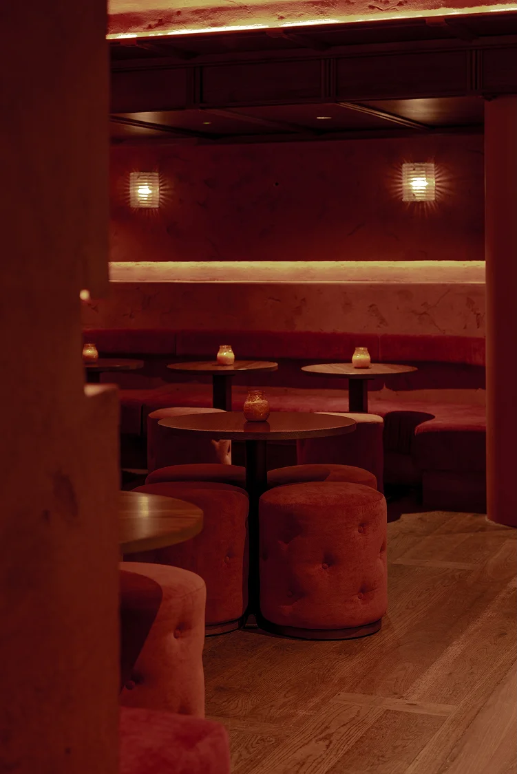 Dimly lit restaurant interior with circular tables and plush seating in shades of red. Cozy ambiance with warm lighting.