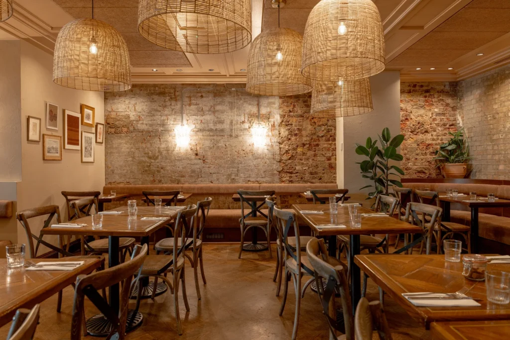 Cozy restaurant interior featuring wooden tables, woven light fixtures, and a brick wall with decorative art.