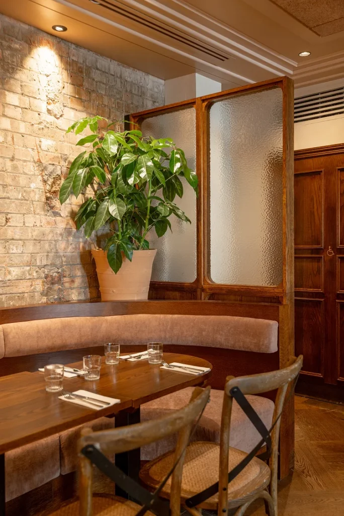 Cozy restaurant interior featuring a wooden booth, glass partition, and potted plant against a brick wall.